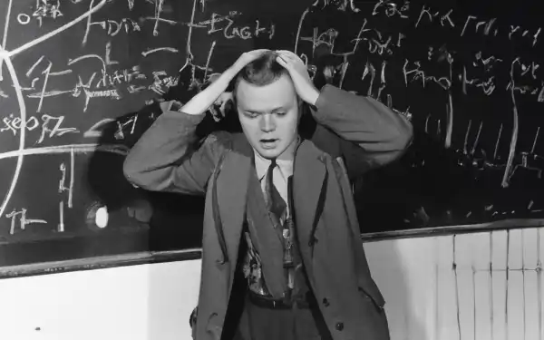 A man in front of a chalkboard scratches his head.